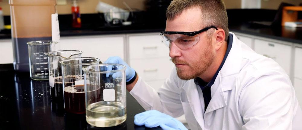 Man wearing safety glasses, blue disposable safety gloves and white lab jacket bends down to countertop to look closely at liquids in test tubes.